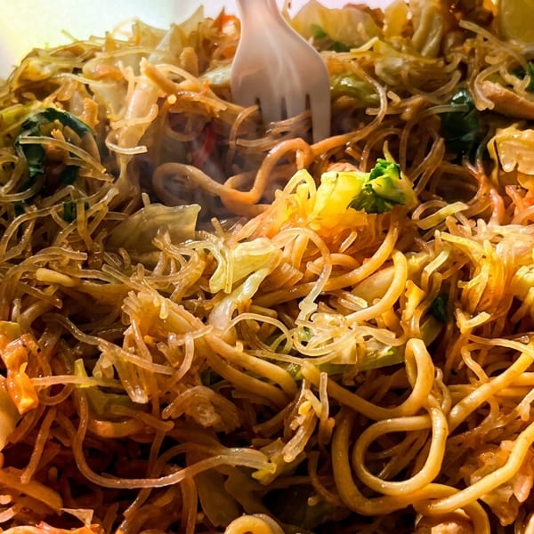 Steaming pancit noodles with vegetables, chicken, and a lemon wedge in a white takeout container.