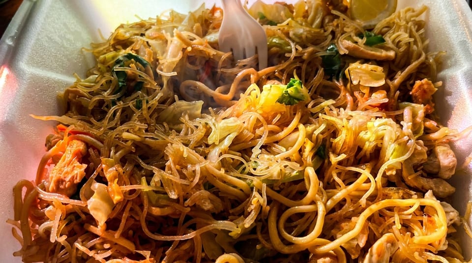 Steaming pancit noodles with vegetables, chicken, and a lemon wedge in a white takeout container.