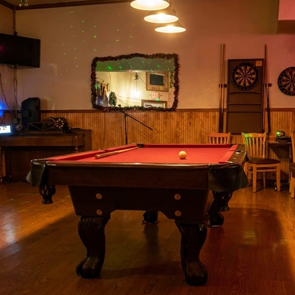 A dimly lit game room with a red-felt pool table, dartboards, and string lights.