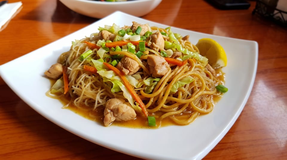 A plate of pancit bihon with chicken, cabbage, carrots, and green onions, garnished with a lemon wedge.